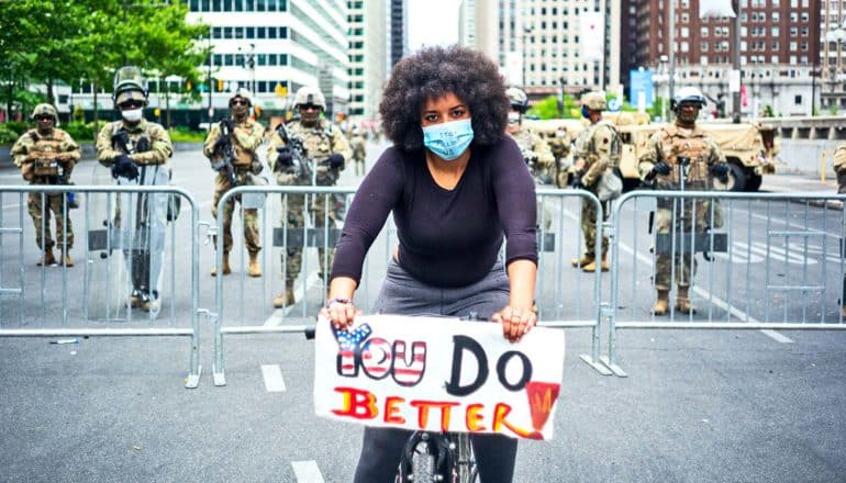 A woman on a bike stands in front of a police barricade holding a sign that reads "You do better" with "you" in the colors of the American flag