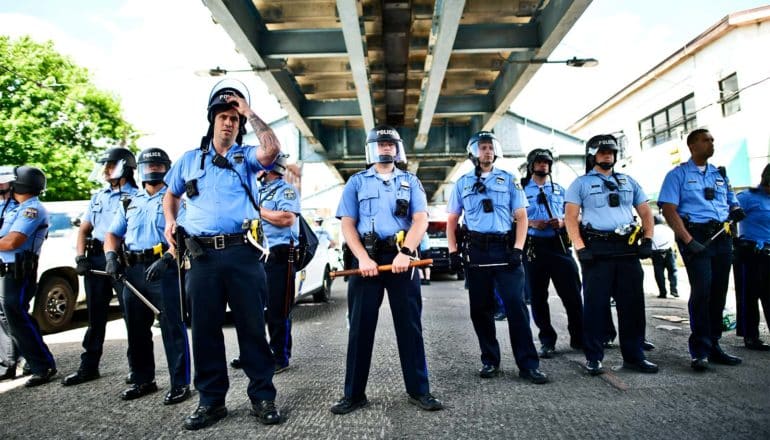 A row of police officers wearing face shields and holding batons stand under a bridge