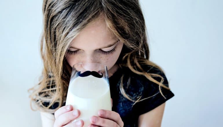 A young girl drinks milk from a glass with a mustache on it