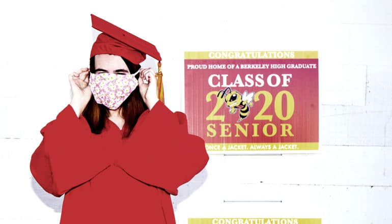 A young woman in a red cap and gown tries to get her face mask on as she stands next to a sign that reads "Class of 2020 senior"