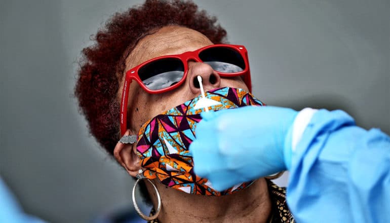 A woman in sunglasses and a colorful mask tilts her head back for a health care worker to collect a sample for a COVID-19 test