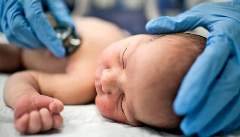 gloved hands hold stethoscope to newborn baby's chest