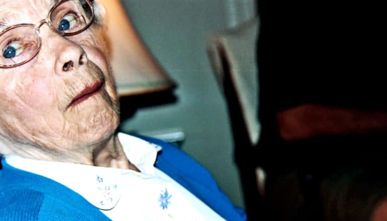 An older woman in a white shirt, blue sweater, and glasses looks over at the camera as if trying to avoid the frame