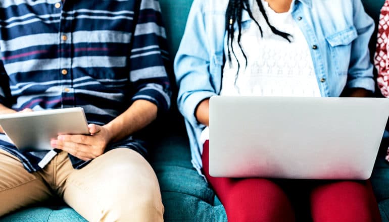 Two people sit on a blue couch, one on a tablet, the other on a laptop