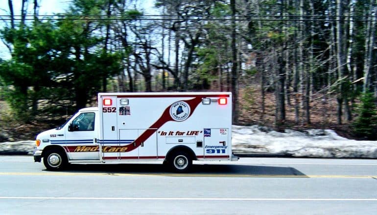 An ambulance drives on a road lined by forest