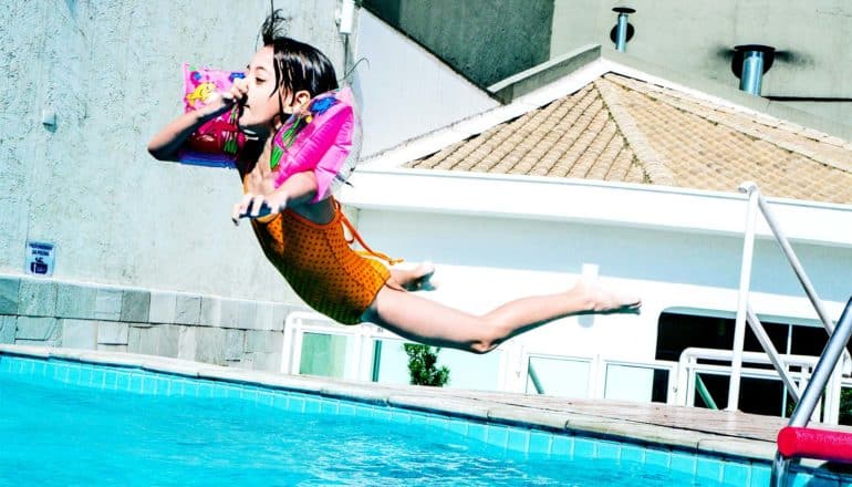 A young girl in an orange bathing suit and pink inflatable floaties on her arms holds her nose as she leaps into a bright blue pool