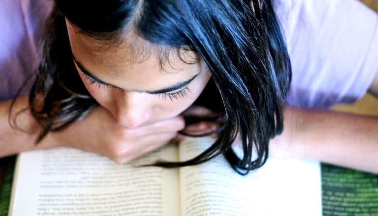 A teen girl is reading a book, resting her chin on her hand on the book