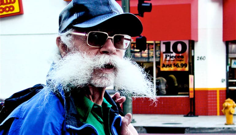 An older man with a large white mustache walks down the street