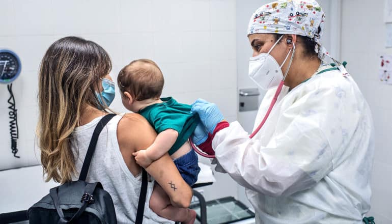 A mother holds her child at the doctor's office as a doctor in full protective gear checks the baby's heart with a stethoscope