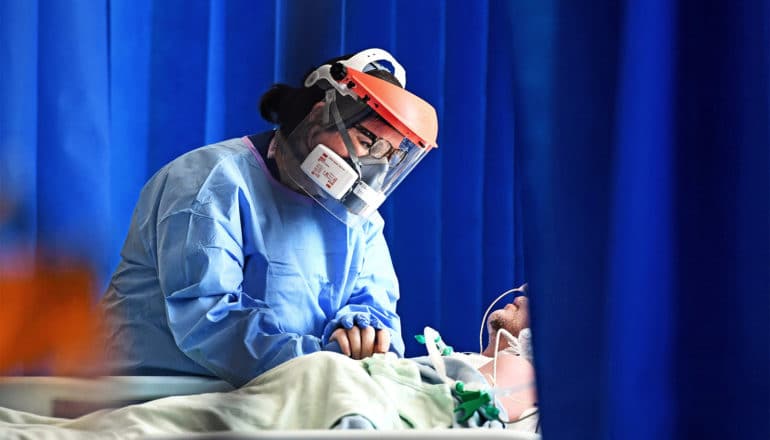 A health care worker in protective masks and gown holds a patient's hand surrounded by blue curtains