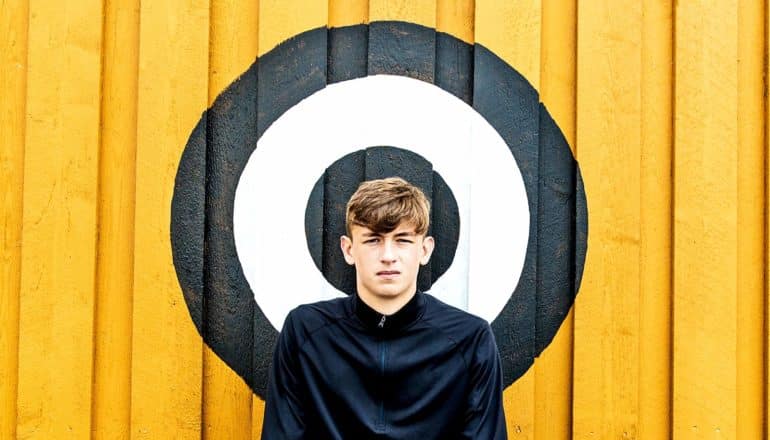 A teenage boy in a black jacket stands in front of a yellow wall with a black and white target on it. His head is in the center of the target.