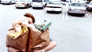 A man carries two paper grocery bags that cover his face through a parking lot