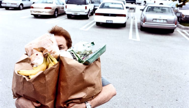 A man carries two paper grocery bags that cover his face through a parking lot