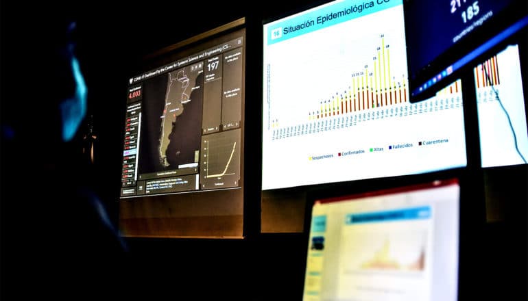 A doctor in a blue face mask looks at COVID-19 statistics on several different computer screens