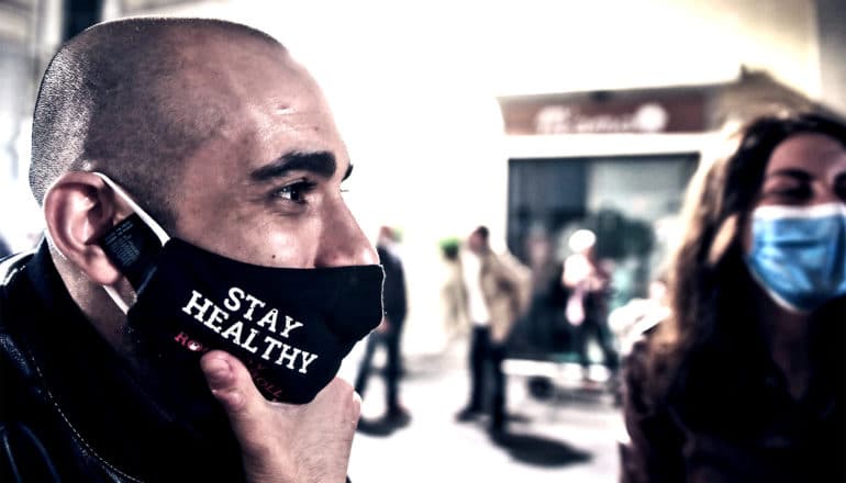 A man in a black mask stands near other people in masks. His mask reads "Stay Healthy"