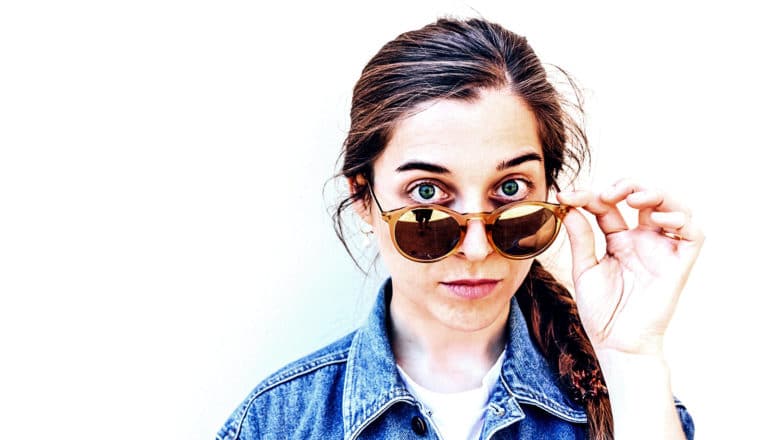 A young woman looks skeptically into the camera, lowering her sunglasses