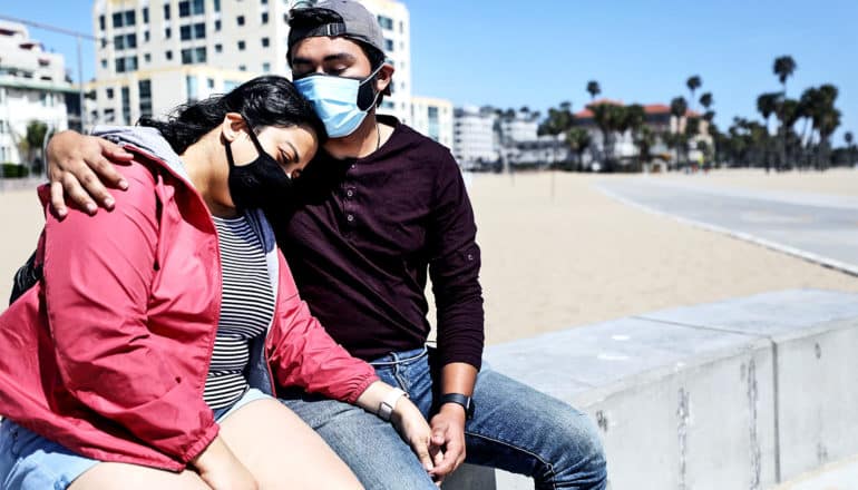 A couple in medical masks sits on concrete on a beach holding each other