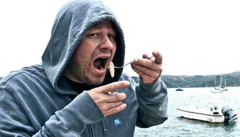 A man looks concerned while raising an oyster on a fork to his mouth