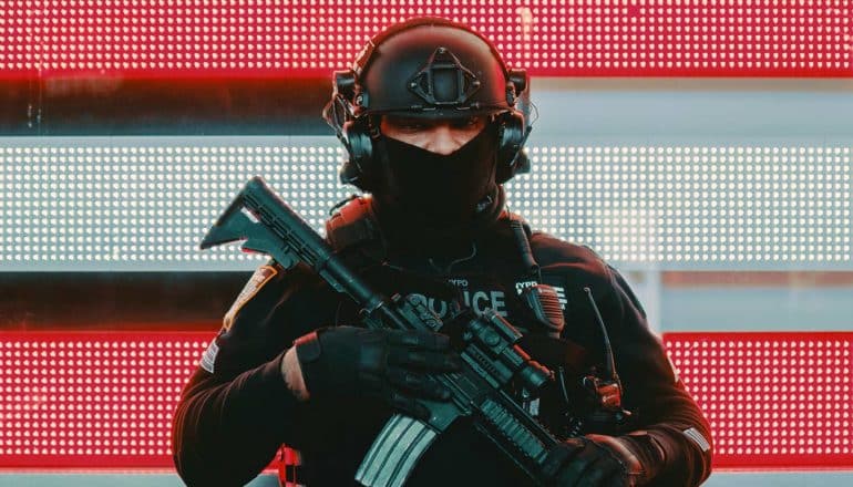 police officer with rifle, mask, helmet, stands in front of light-up American flag