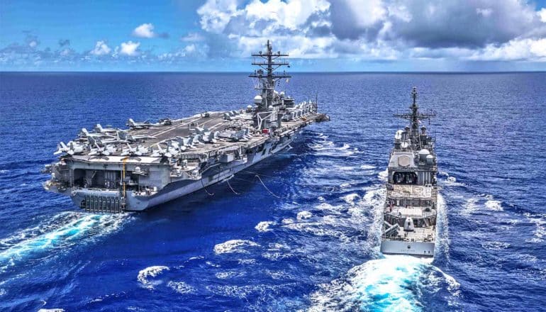 A smaller ship moves through the ocean next to a much larger aircraft carrier