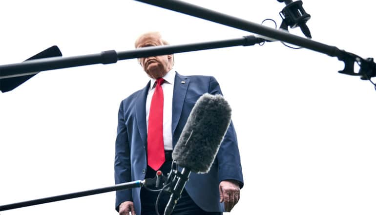 President Trump stands against a white sky with microphones criss-crossing the frame