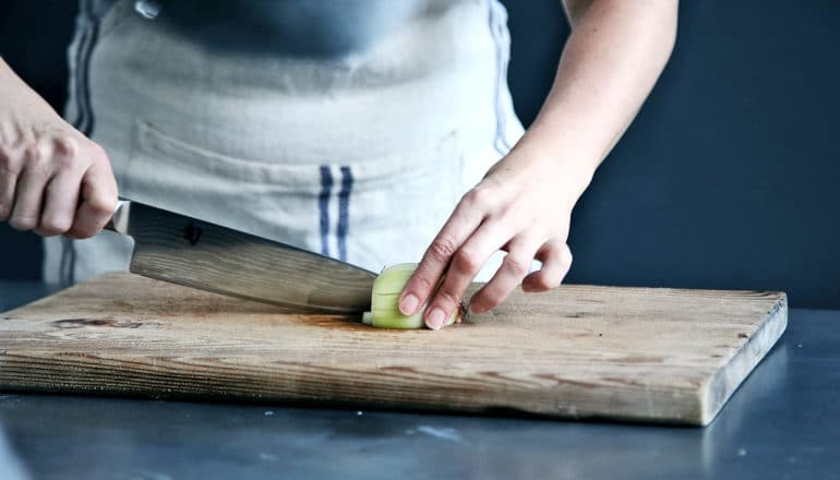 hands cut onion with knife on wood cutting board on metal counter