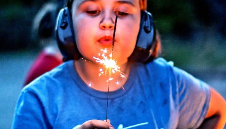 A young girl wearing a blue shirt and black headphones blows on a sparkler