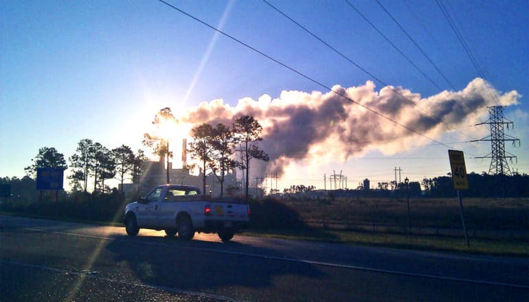 A pickup truck drives past a factor emitting plumes of smoke against a blue sky