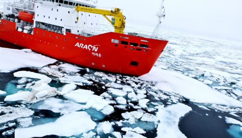 A ship with a red hull moves through sea ice in the Arctic Ocean