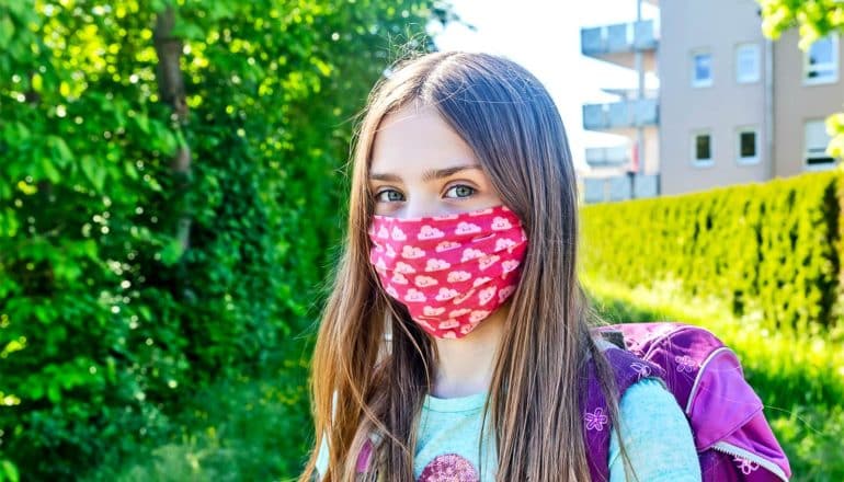 A young girl wears a red mask and a backpack in a green yard