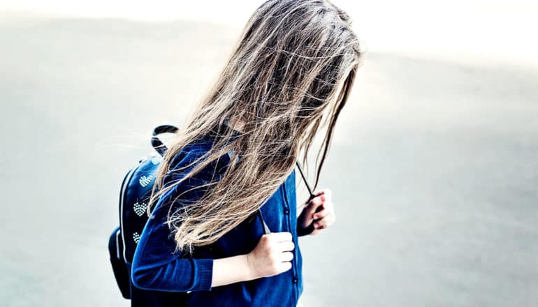 A young girl walks alone wearing a backpack and looking down at the road