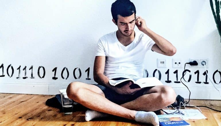 A college student sits against a wall while charging his smartphone and reading a text book