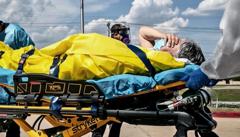 A person on a stretcher holds their hand over their face in pain as first responders take them into an ambulance