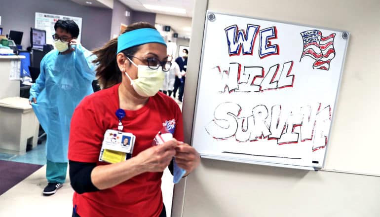 A health care worker wearing a face mask rushes past a message on a whiteboard that reads, "We Will Survive" with the American flag next to it