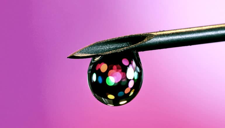 A drop of liquid falls from a syringe, with colored light shining through it