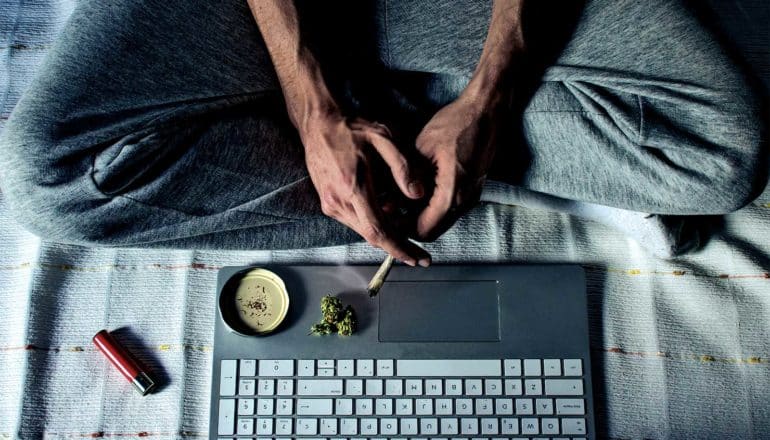 A man holds a joint over his computer as he sits on a bed with legs folded looking at the screen