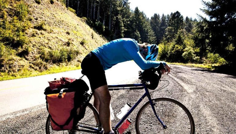 A man on a bicycle pauses by the side of a mountain road and leans down to rest on the bike's handlebars