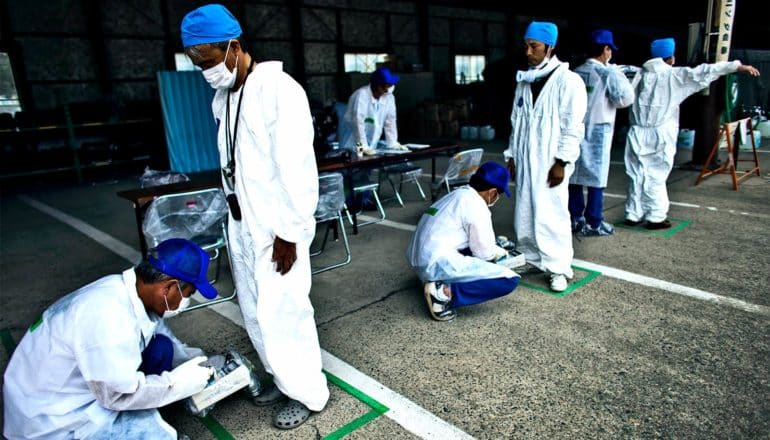 A line of people are scanned for signs of radiation while wearing white protective suits and face masks