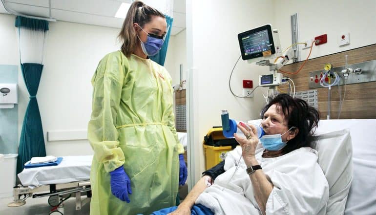 A health worker stands over a patient in bed breathing into a tube