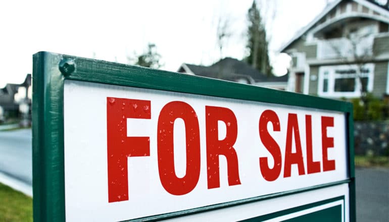 A for sale sign stands next to a suburban street