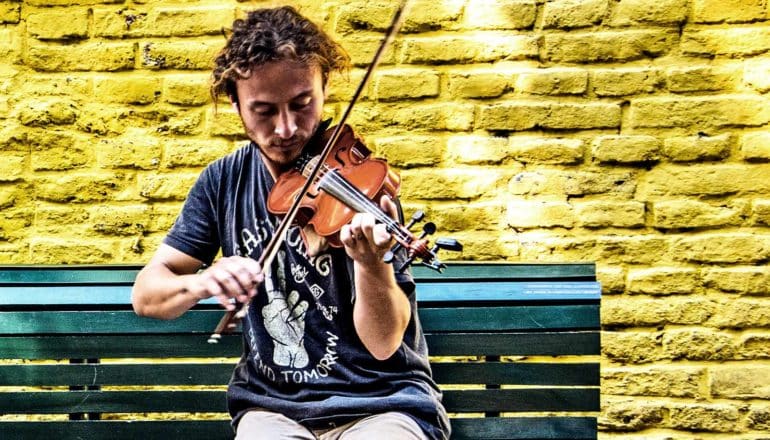 A man plays a violin while sitting on a bench near a yellow wall