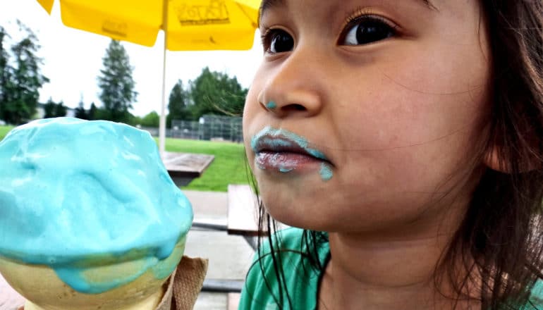 A little girl eats blue ice cream, and has some around her mouth and on her nose