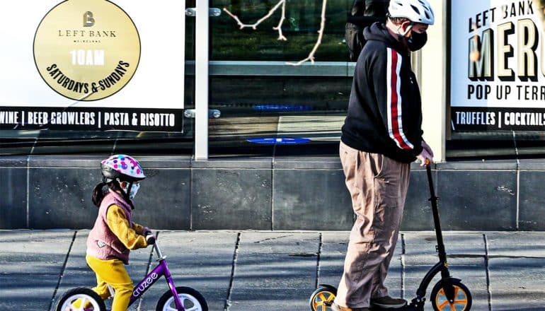 A father wearing a face mask rides a scooter ahead of his young daughter, also wearing a mask, on her bike