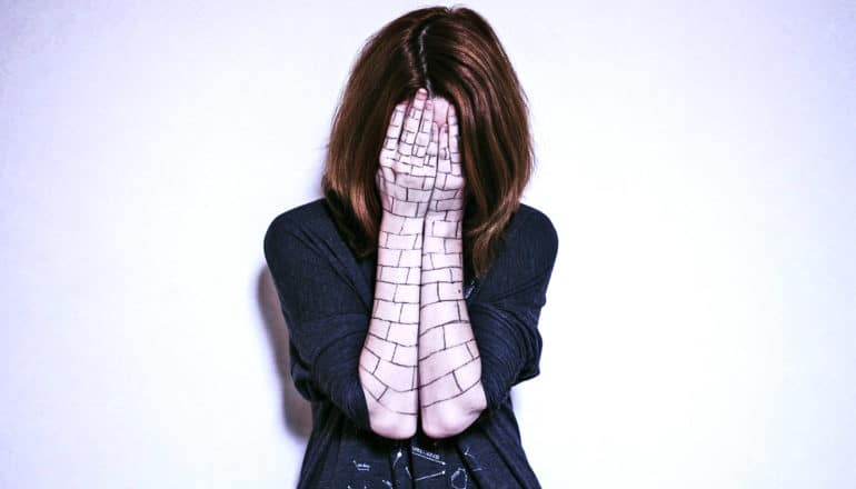 A woman puts her face in her hands. Her arms are covered in lines that make her arms look like a brick wall