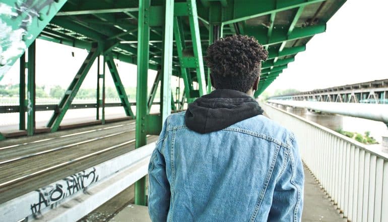 A young man in a jean jacket walks on a sidewalk under a green bridge