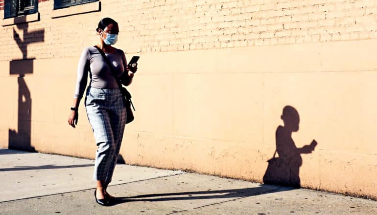 A woman walks down the street wearing a mask and looking at her phone