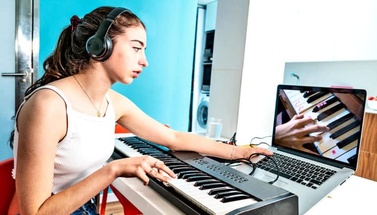 A young woman plays piano with one hand while holding up a laptop screen showing a hand on piano keys with another