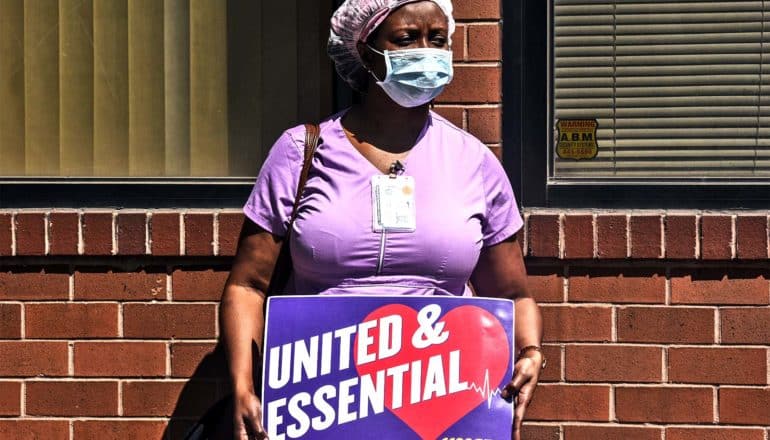 A health worker in purple scrubs protests shortages at nursing homes, holding a sign that reads "United and Essential"