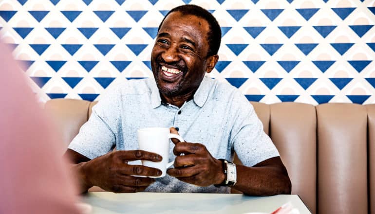 An older man sits in a booth with a cup of coffee as he smiles at the person across the table