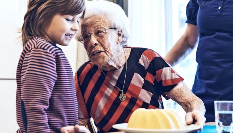 A grandmother leans over to speak to her grandson while holding a plate with a cake on it.